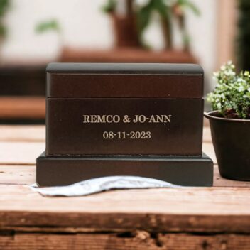 Groot donker gebeitst houten ringdoosje (10x7x7 cm) met persoonlijke gravure van namen en trouwdatum in een rustieke setting.
