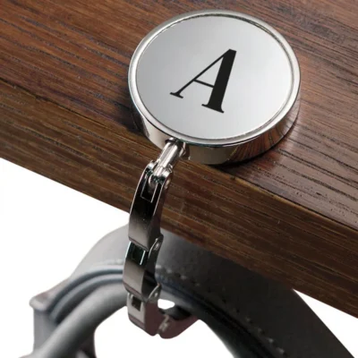 Zilverkleurige tas haak met initiaal in gebruik op tafel: hangt een tas veilig en stijlvol aan de rand, perfect als persoonlijk cadeau voor haar