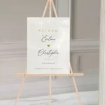 Gepersonaliseerd welkomstbord Love Heart Edition op wit acryl met sierlijk kalligrafie-schrift en gouden hartje op houten ezel