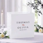 Witte acryl Romeo enveloppendoos met namen Thomas en Olivia in kapitalen, twee blush hartjes en datum 26.06.2026, op receptietafel met witte rozen en brandende kaars