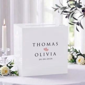 Witte acryl Romeo enveloppendoos met namen Thomas en Olivia in kapitalen, twee blush hartjes en datum 26.06.2026, op receptietafel met witte rozen en brandende kaars